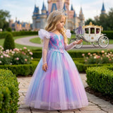 A young girl stands in a garden wearing a princess-style tulle dress with a blue, pink, and purple iridescent gradient, sheer long sleeves, puffed shoulders, and a matching tiara. A castle and carriage are visible in the background.